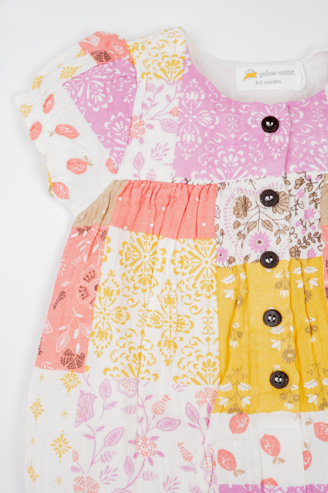 Baby romper in soft organic cotton with colourful patchwork print in pink, yellow and white tones, featuring floral and geometric patterns. Designed with snap buttons for easy dressing and diaper changes.
